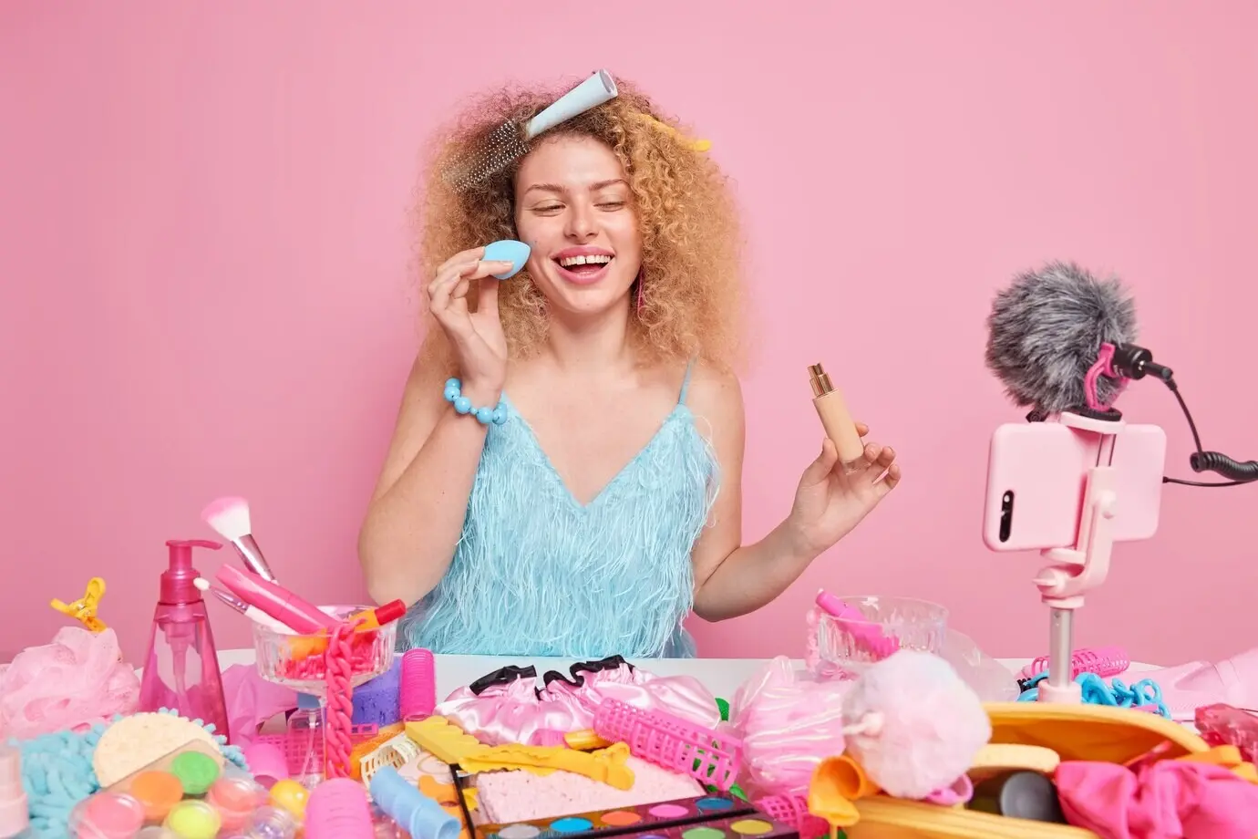 A woman blogger in a blue dress applies foundation to her face while a comb is stuck in her hair, sitting at a table surrounded by beauty products and using a mobile phone to record a video blog.