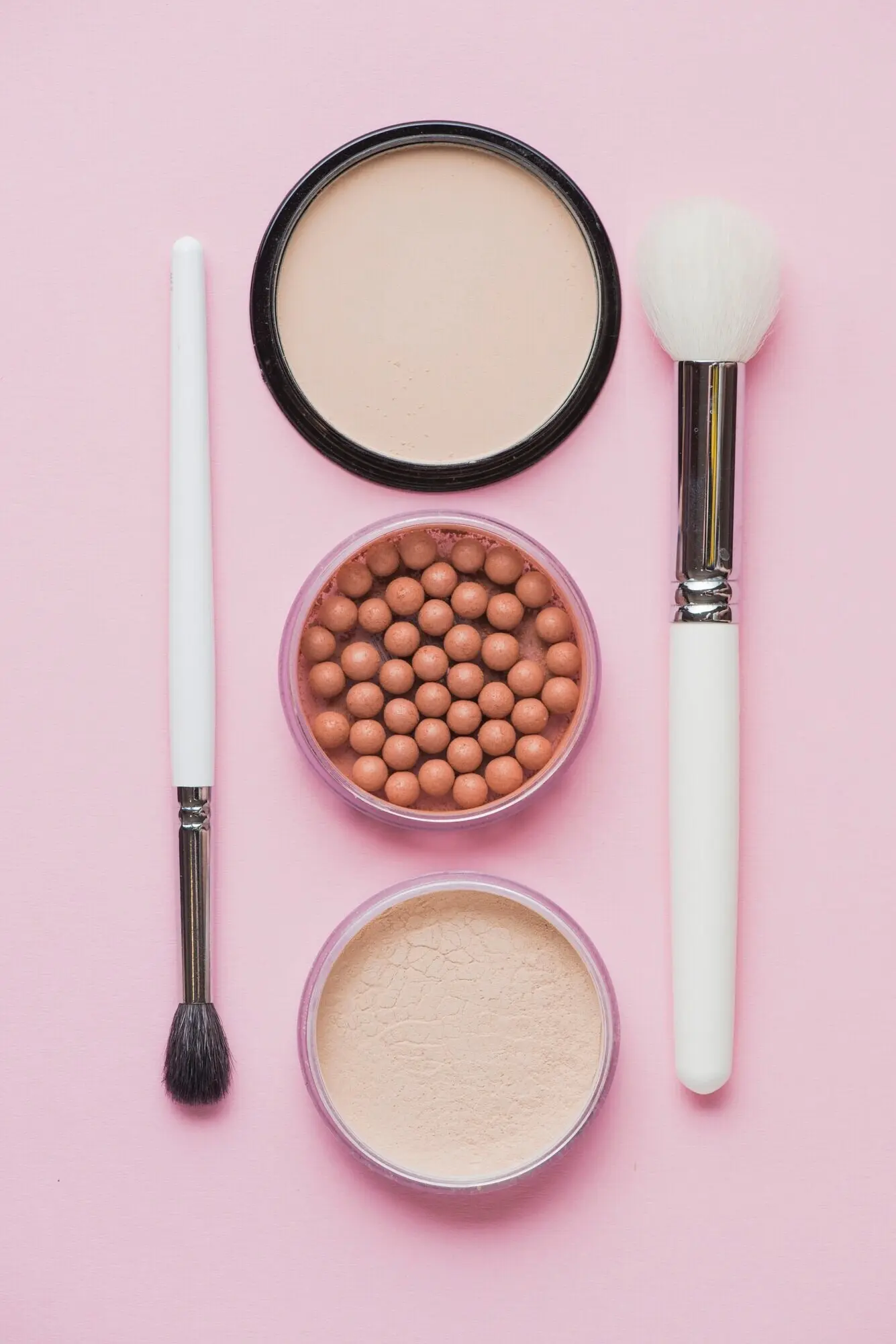 Compact powders, bronzing pearls, and makeup brushes on a pink surface.